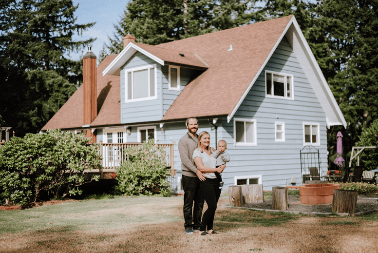 Jason and Haley with their Newborn baby, standing in front of a house. The house is white in colour, and has a classical look about it.
