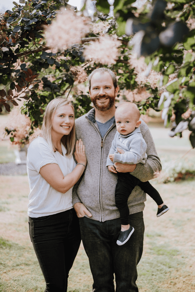 Jason and Haley standing in a field with their Newborn baby, there is a tree in the background, and some foliage in the foreground.